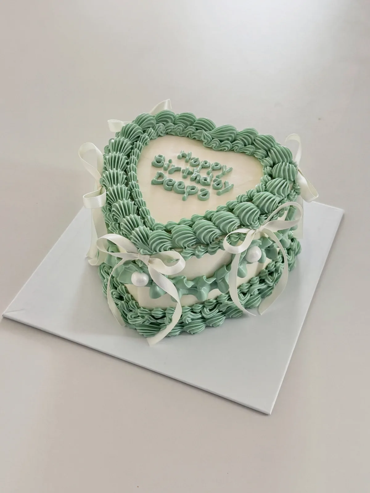 Sage green heart cake with white bows and shell piping, handmade to order by Danine Cakes in Streatham, South London