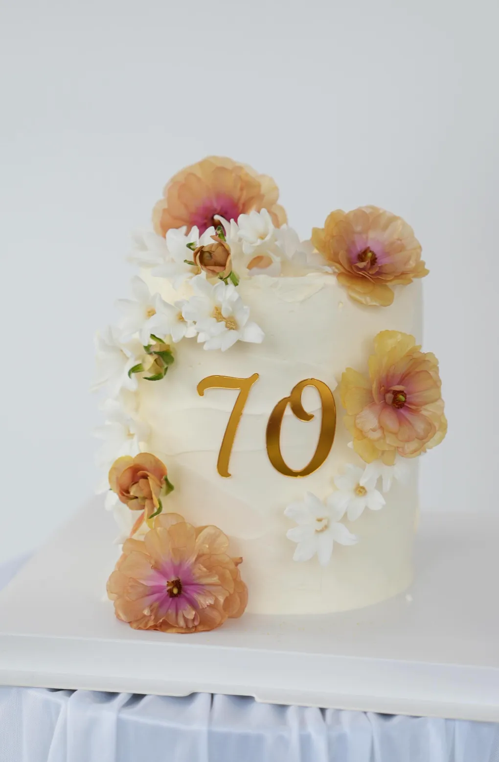 White 70th birthday cake decorated with fresh peach ranunculus and white blossom